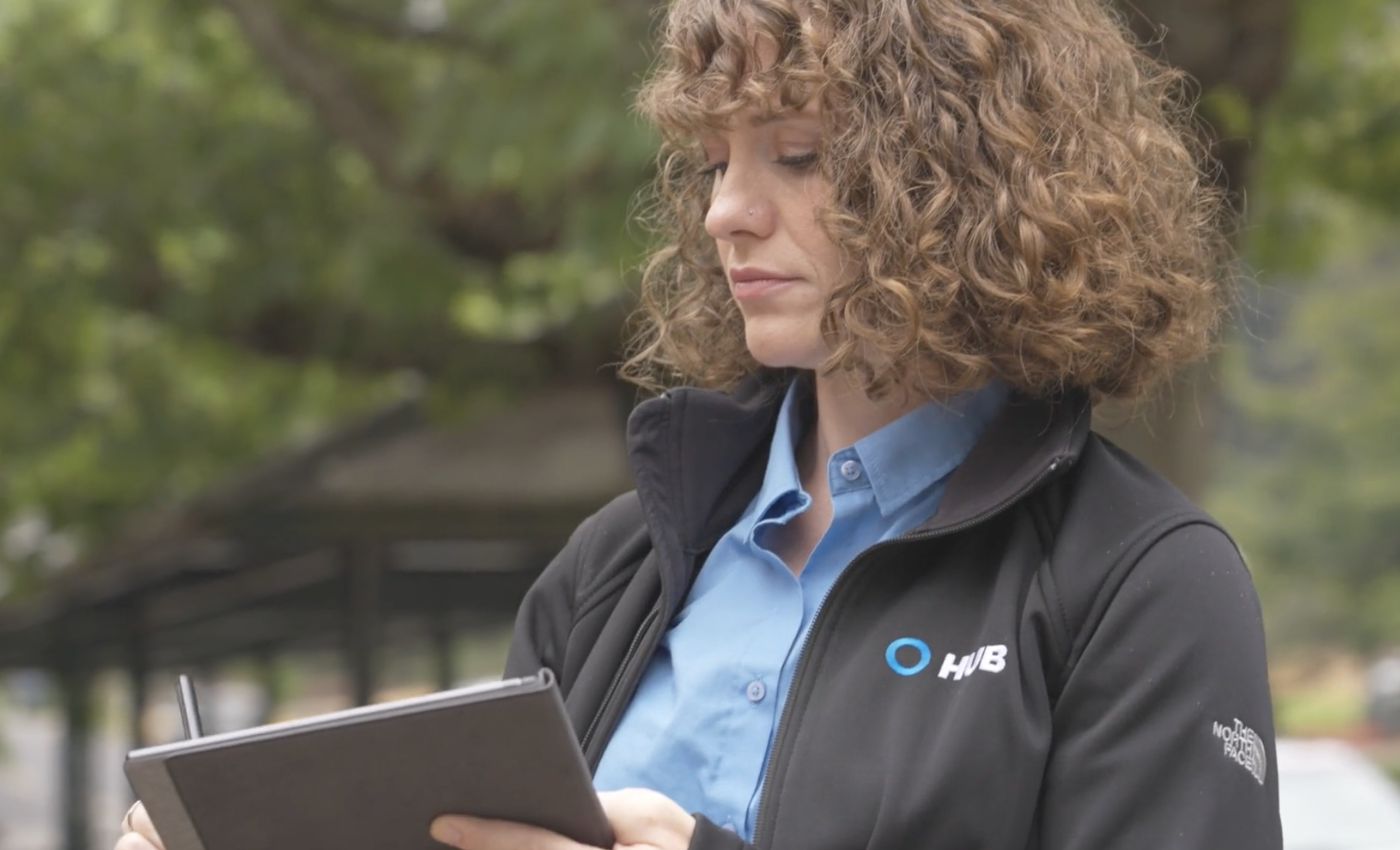 A HUB International commercial lines advisor reviewing an account on a tablet outdoors, HUB-branded jacket visible.