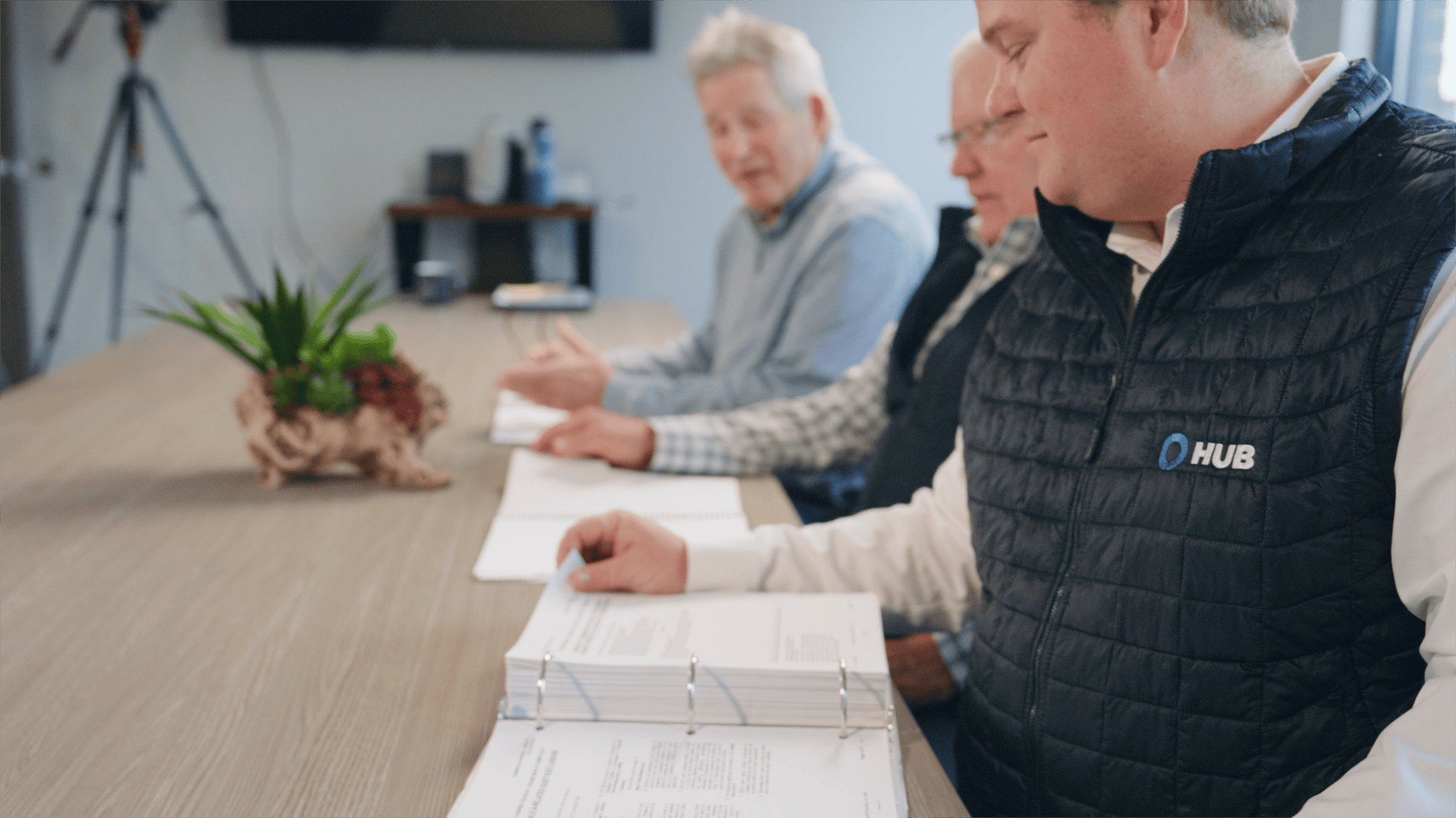 Three Team Haugen principals reviewing a client program at a conference table, HUB International vest visible in the foreground.